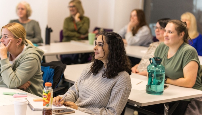 People seated at tables in a bright classroom setting with notebooks and refreshments. The central person in a grey ribbed jumper looks particularly engaged and attentive, smiling slightly while looking towards the front of the room, with other learners visible in the background.